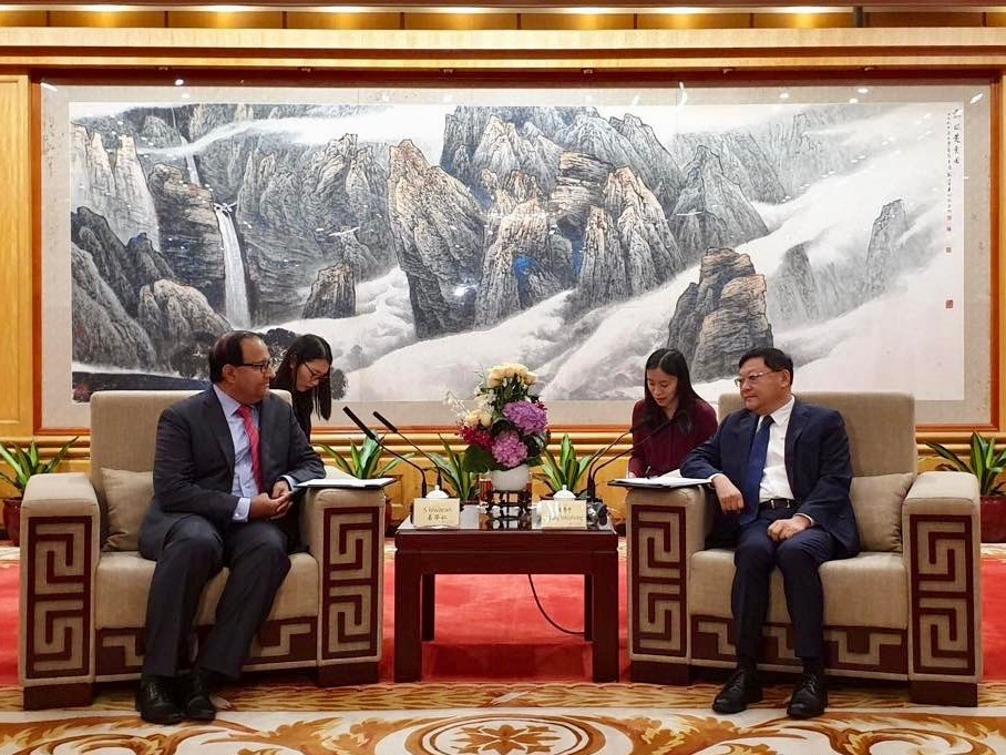 Two suited men seated in ornate chairs across from each other, with two women behind them. Landscape painting above.